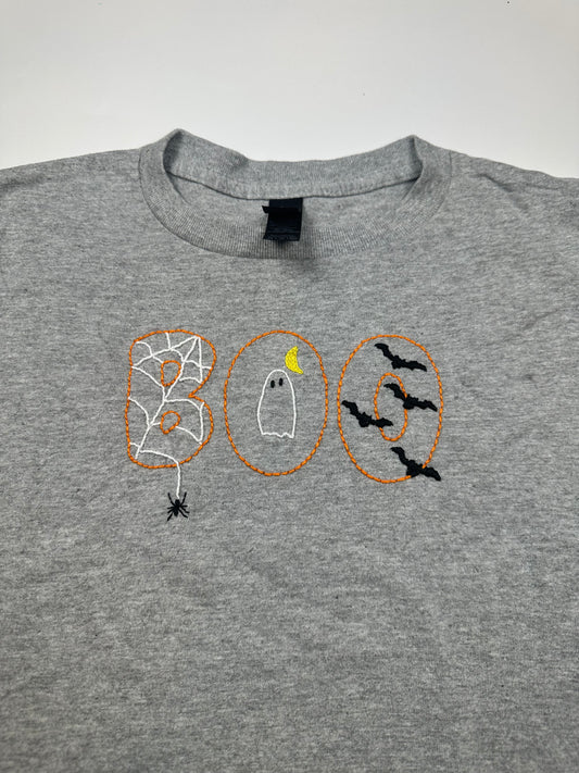 A heather gray crewneck t-shirt featuring a 'BOO' Halloween graphic across the chest. The letter 'B' is filled with a spiderweb outline and a black spider, 'O' in the middle features a white ghost with a yellow crescent moon, and the letter 'O' is outlined with black bats.